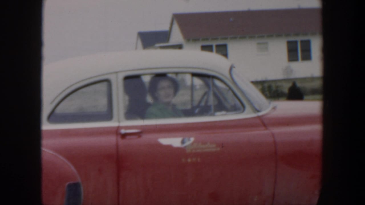 Woman seated in a red-and-white classic car in front of houses.