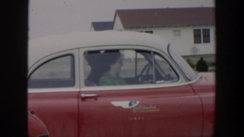 A woman in a dark hat and green top sits inside the red-and-white classic car, with residential buildings in the background.