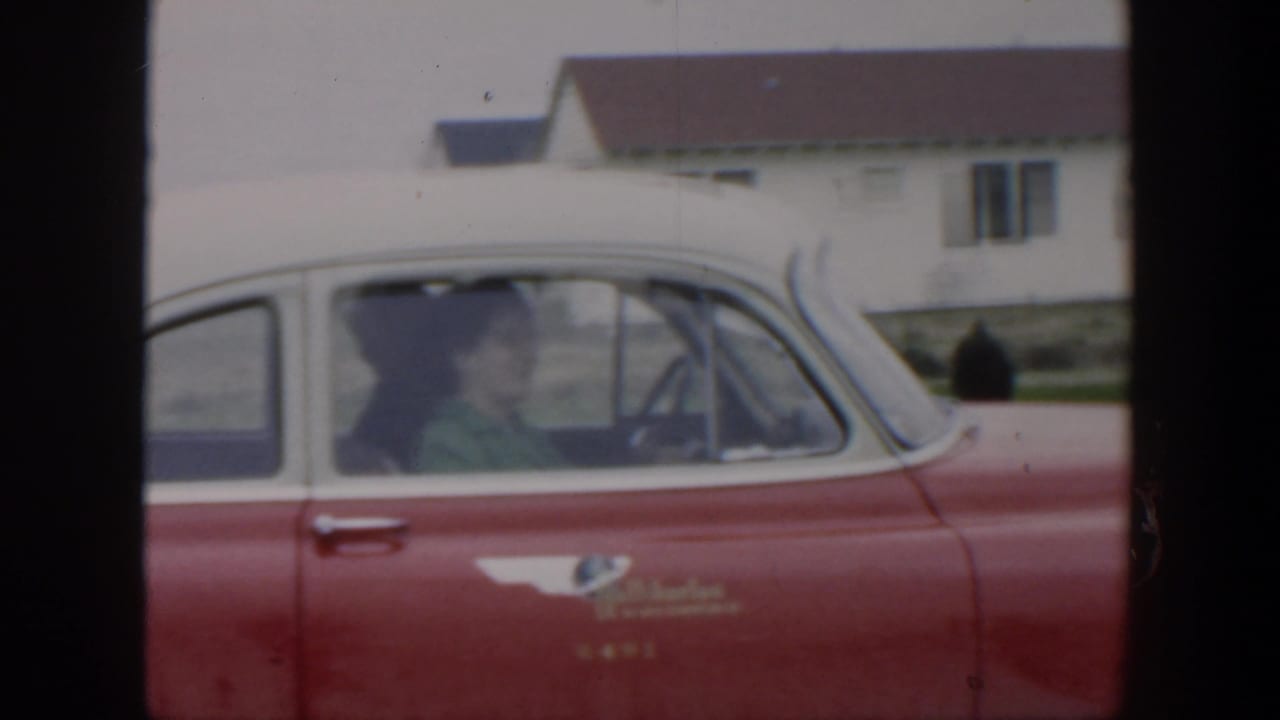 Red and white vintage car beside a simple house, with a woman visible through the passenger window.