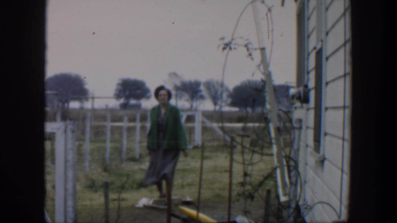 Woman in a green cardigan walking beside a white fence in a rural yard.