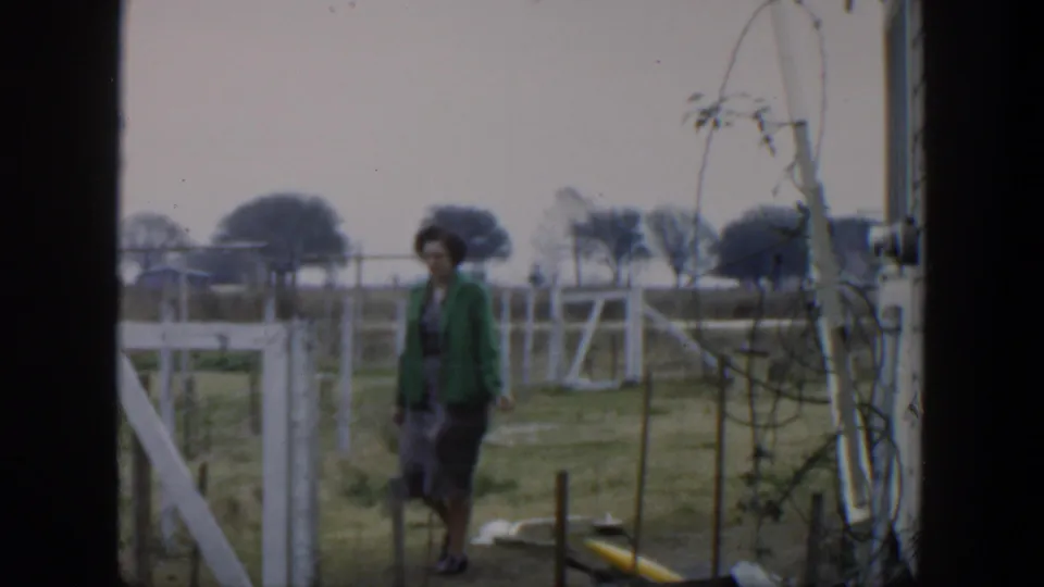 The woman moves carefully through the rural yard, with white fence rails, tangled wires, and open land visible behind her.