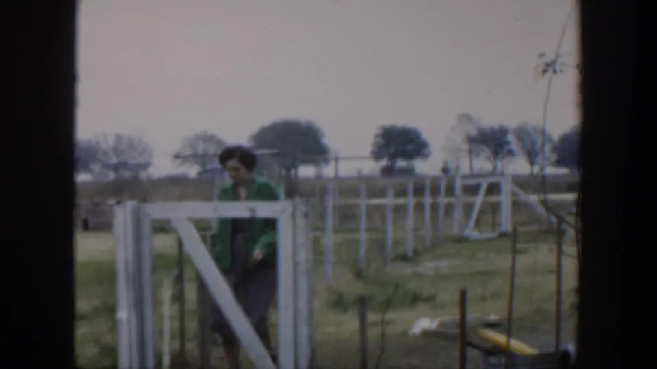In the Cuba home-movie scene, the woman passes behind a white gate or fence section with grass, wires, and trees in the background.
