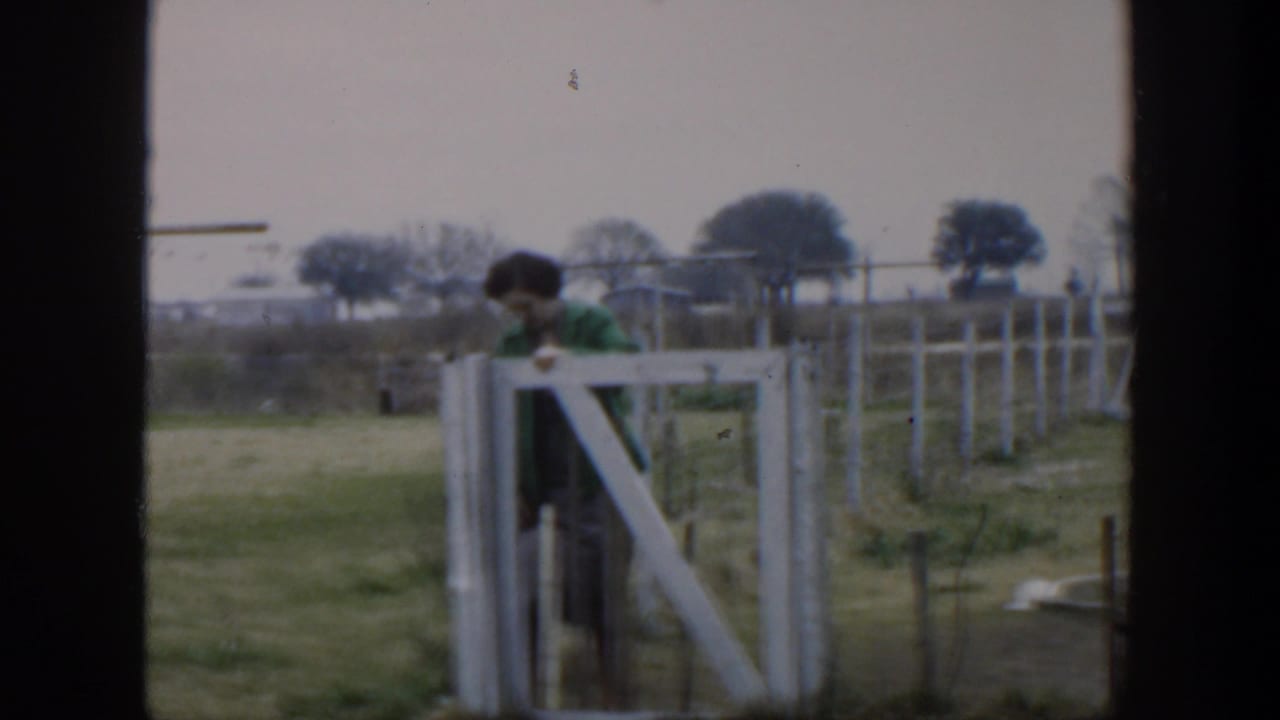 1951 Cuba home-movie frame showing a woman in a green jacket leaning near a white wooden gate in a fenced rural area.