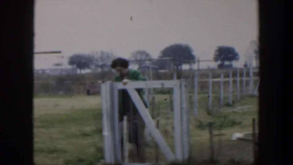 1951 Cuba home-movie frame showing a woman in a green jacket leaning near a white wooden gate in a fenced rural area.