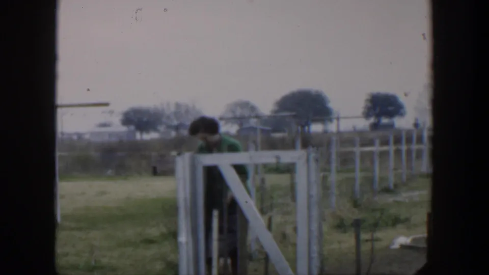 1951 Cuba rural frame with a woman in green at a white gate, wire fence lines, grassy ground, and distant trees under gray sky.