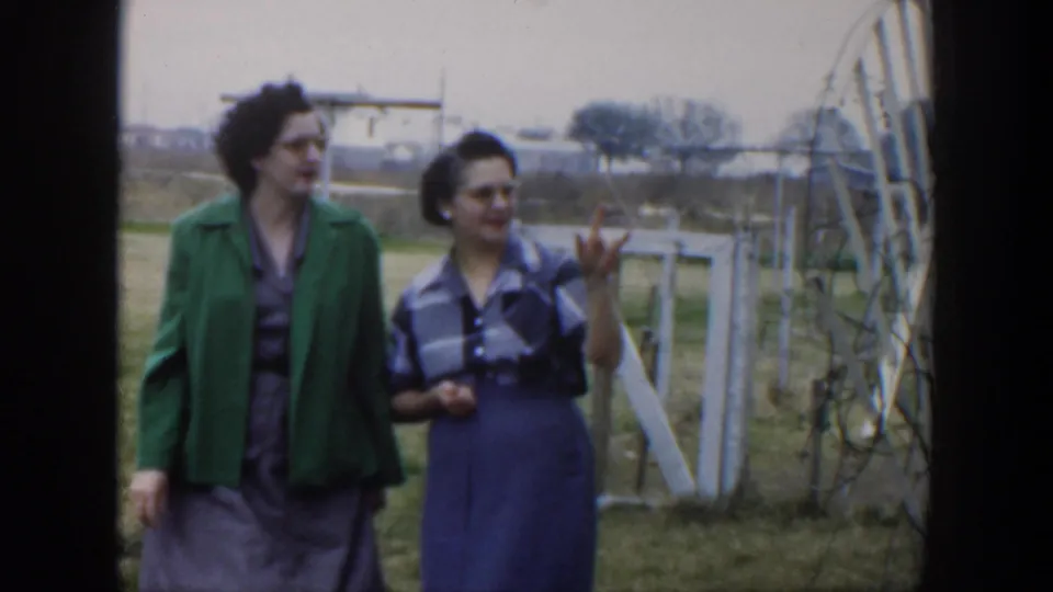 1951 Cuba home-movie frame showing two women walking near a white gate, with one gesturing beside fencing and open grass.