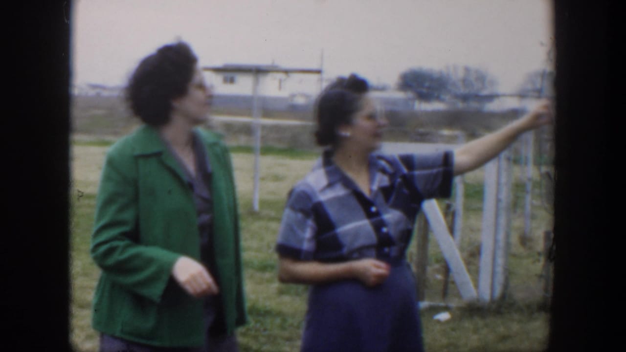 Two women stand in a grassy field beside a wooden fence.