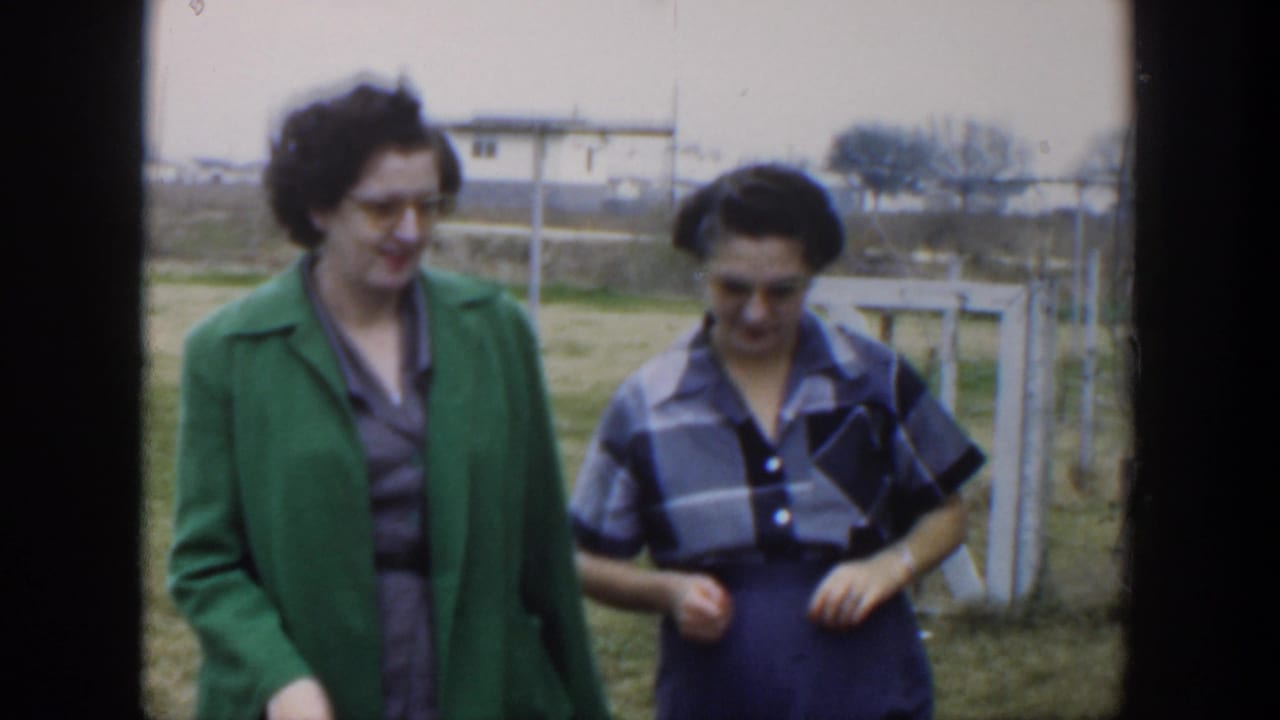 Two women walk near a fence with grass and buildings behind them.