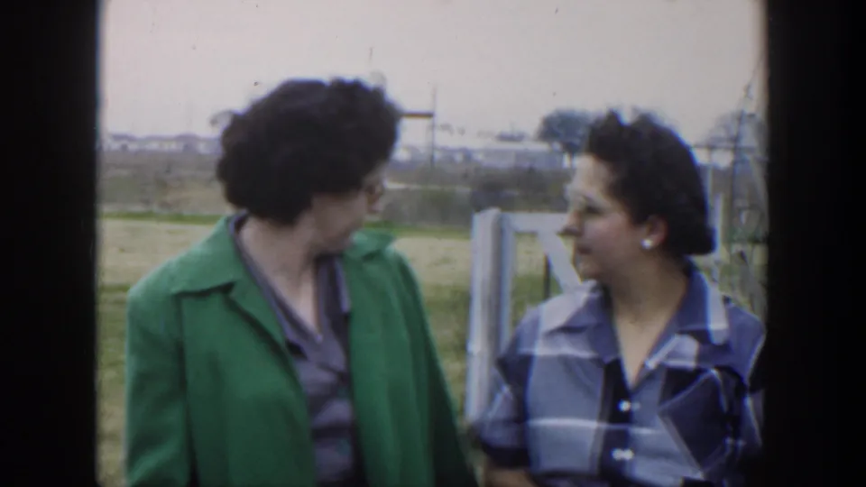 Two women in 1951 Cuba face each other while talking beside a wooden fence in a grassy rural setting.