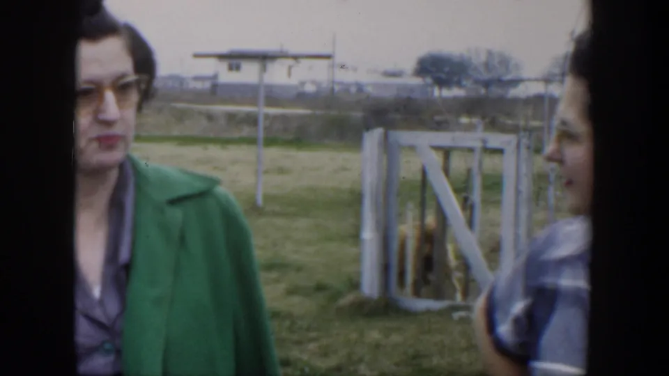 A woman in a green jacket stands near a rural fence in Cuba, with the second woman partly visible at the edge of frame.