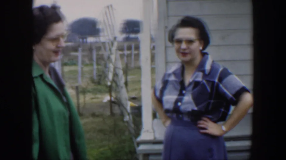In metadata-identified 1951 Cuba, two adult women in glasses stand near house siding and porch posts during a casual outdoor conversation.