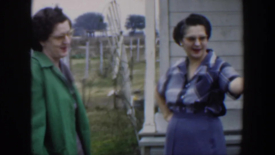 Two adult women in 1950s attire appear in conversation outside a residence, with one woman gesturing near the frame edge.
