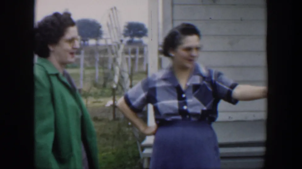 In this 1950s home-movie frame, two adult women stand near a porch as the woman in plaid gestures toward someone off camera.