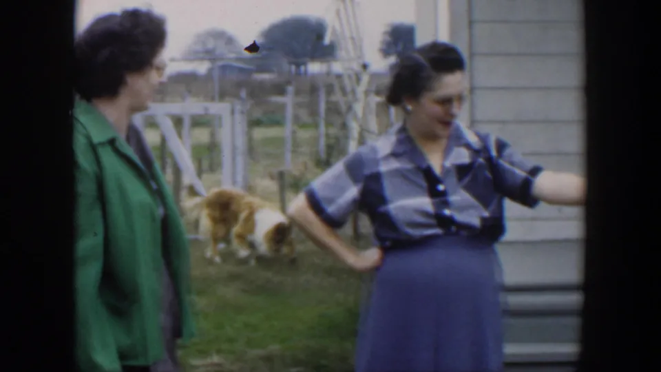 Two adult women in metadata-identified 1951 Cuba stand outside a residence, one gesturing while a dog moves through the yard behind them.