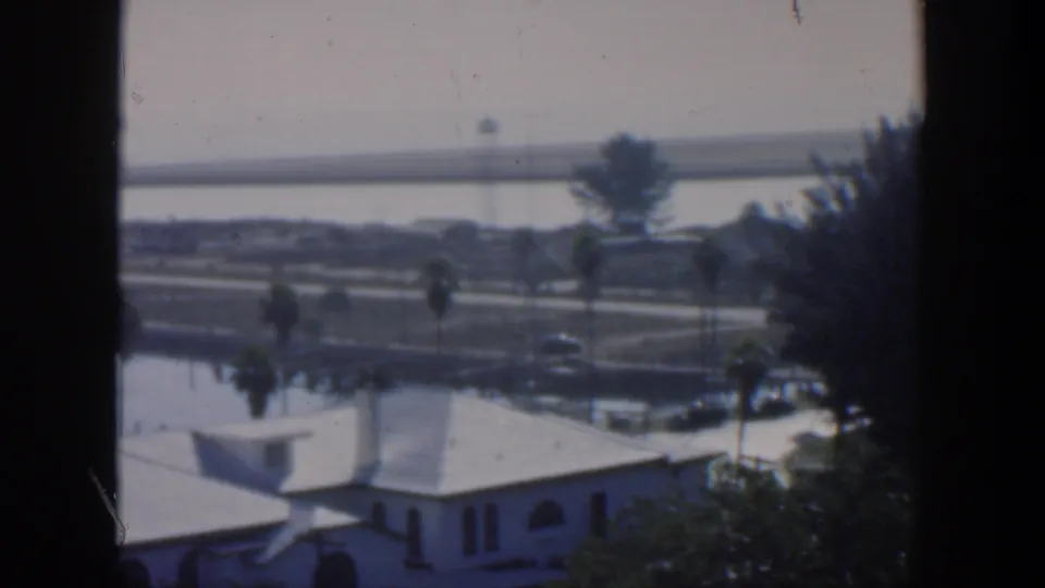 1951 Cuba frame showing a palm-lined waterfront with low white buildings, roadways, water, and dark window-like foreground edges.