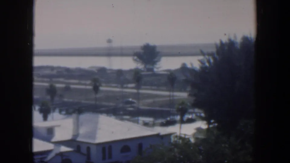 1951 Cuba home-movie frame looking from a window over white low-rise buildings, palm trees, a road, water, and a far shoreline.