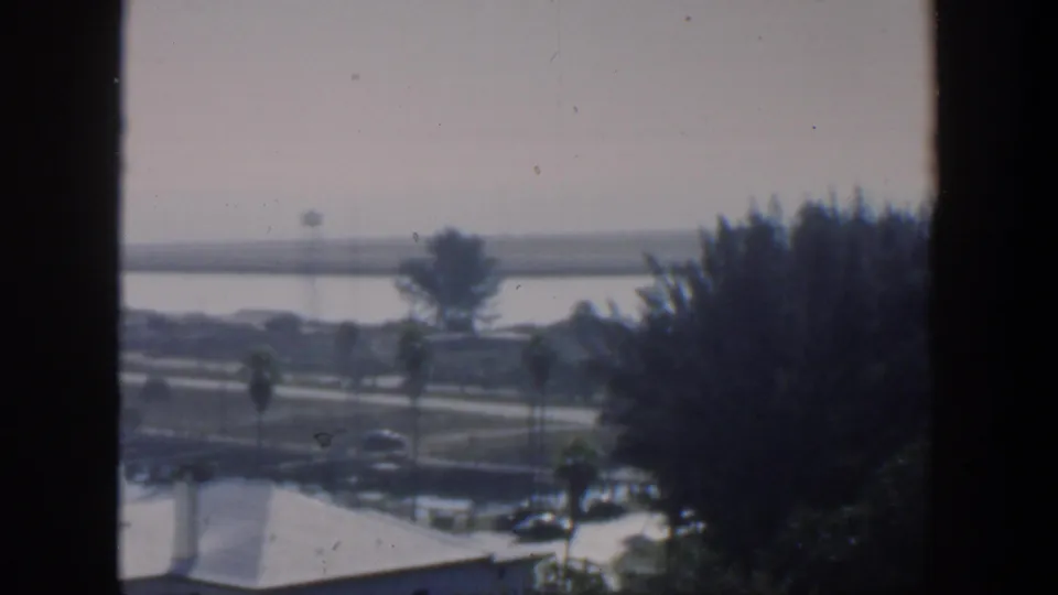 1951 Cuba exterior view through a dark window frame, with palms, low white buildings, parked cars, water, and a distant shore.