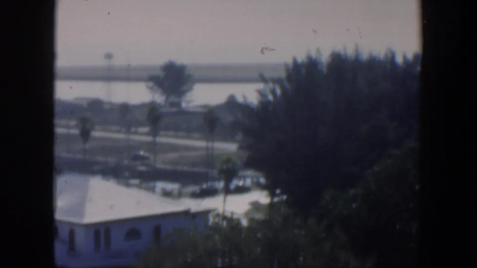 Soft 1951 Cuba home-movie frame showing white stucco buildings, palms, a waterfront road, water, and distant land beyond.