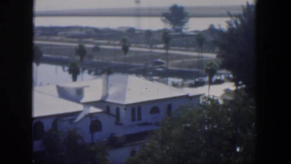1951 Cuba frame with a white building in the foreground, palms and cars near a road, and water with distant shoreline behind.