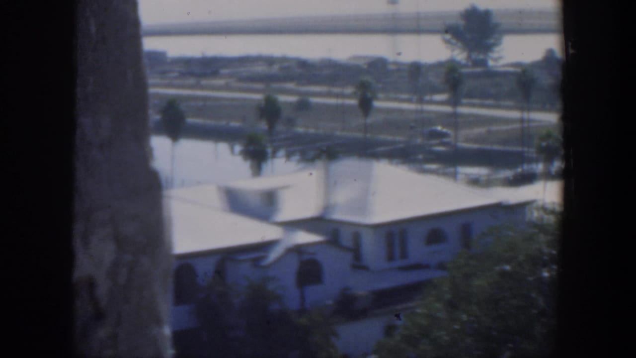 1951 Cuba 8mm frame showing a white stucco house below, palms, a waterfront road, calm water and a distant bridge from an elevated viewpoint.