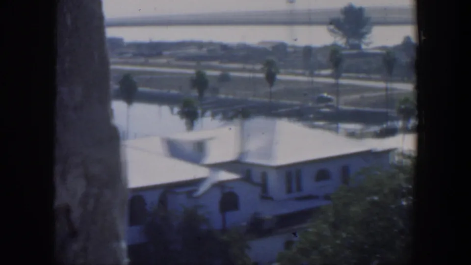 1951 Cuba 8mm frame showing a white stucco house below, palms, a waterfront road, calm water and a distant bridge from an elevated viewpoint.