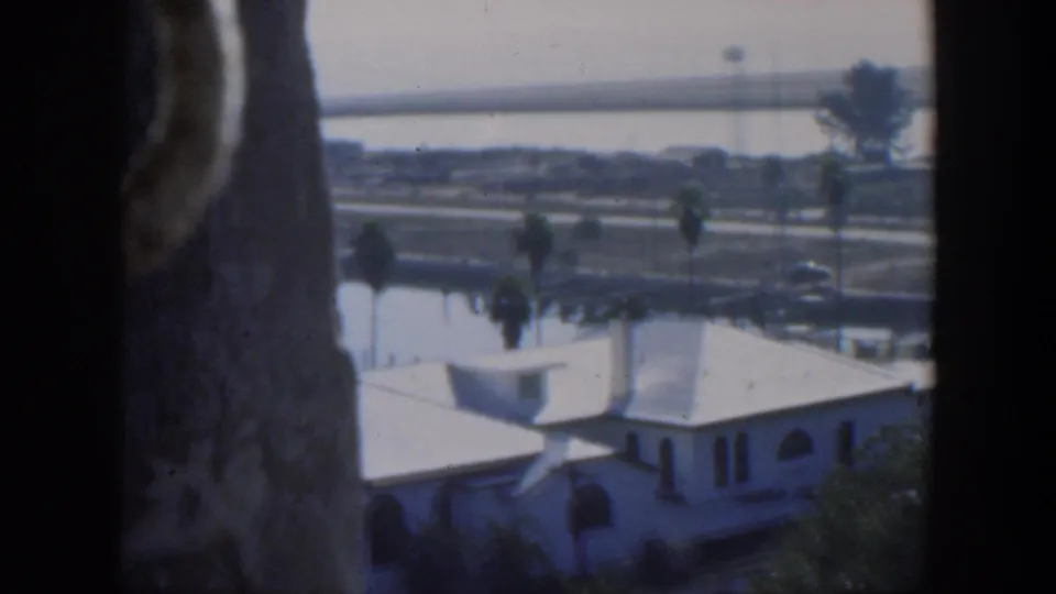 1951 Cuba home-movie frame with a white house partly blocked by dark framing, palm-lined water, road traffic and a distant bridge.