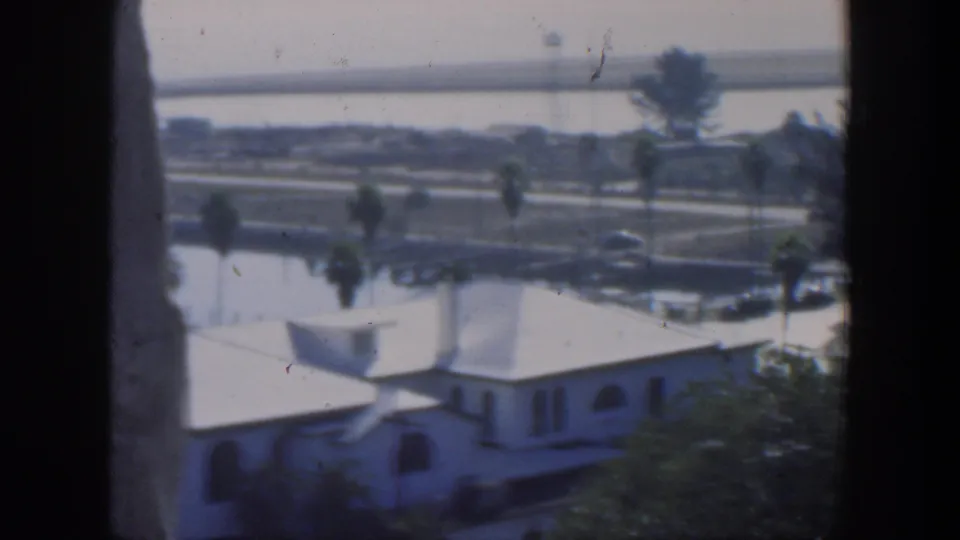 1951 Cuba 8mm still showing the house lower in frame with palms, a waterside road, small cars, calm water and bridge beyond.