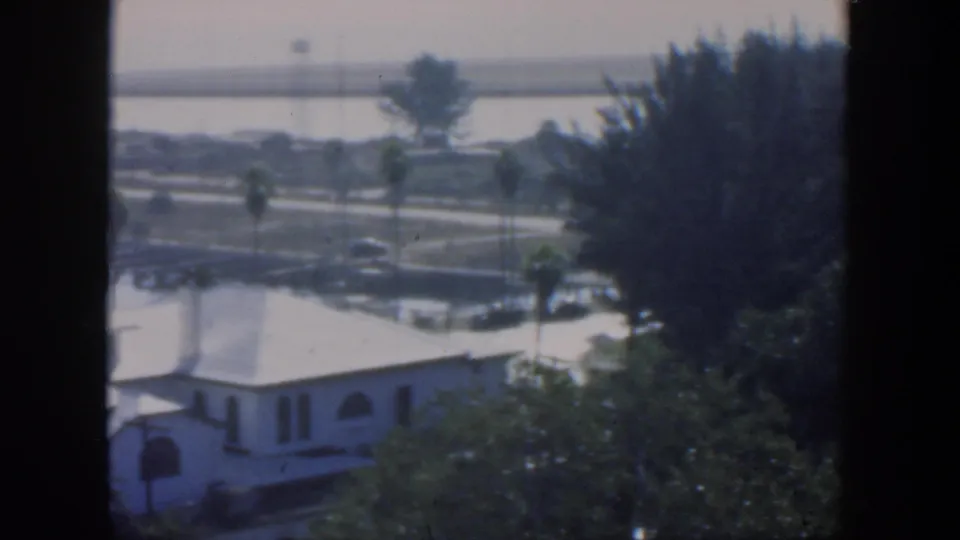 1951 Cuba frame with a white residential building, trees in the foreground, palms along the road, calm water and a distant bridge.