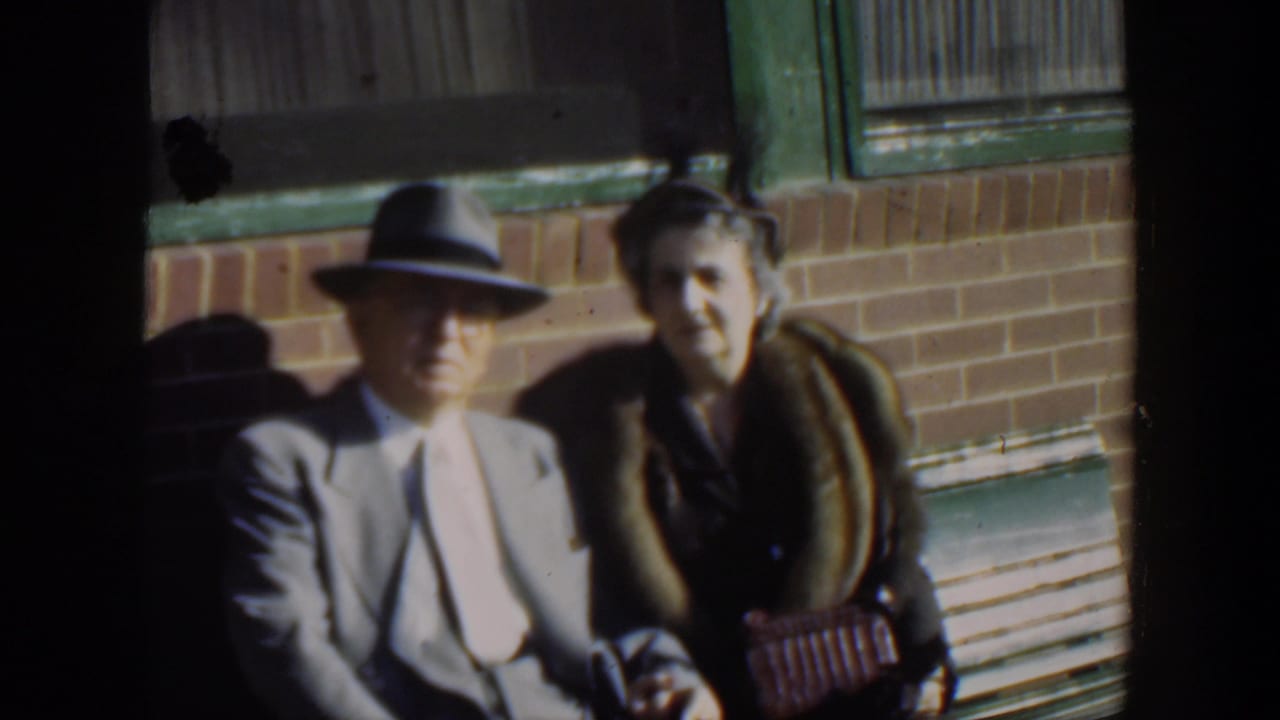 Man in a fedora and woman in a fur-collar coat sit together outside a brick building.