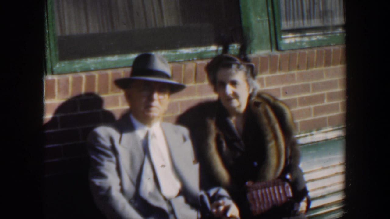 In 1951 Cuba home-movie footage, an older couple sits close together on a bench in formal clothing outside a brick building.