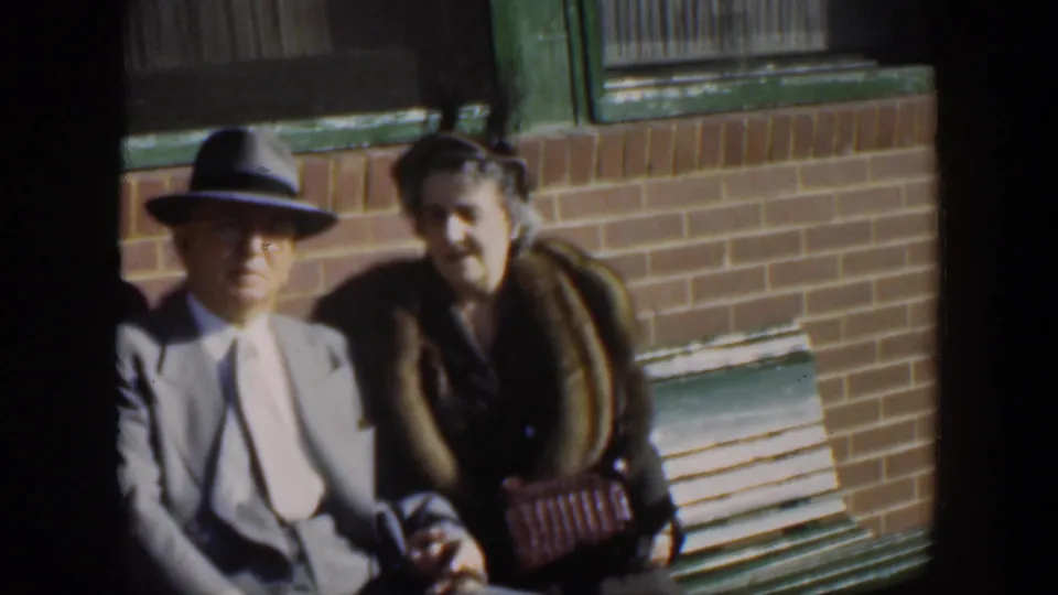 The couple remains seated in bright sunlight, with the man in a light suit and fedora and the woman holding a woven purse.