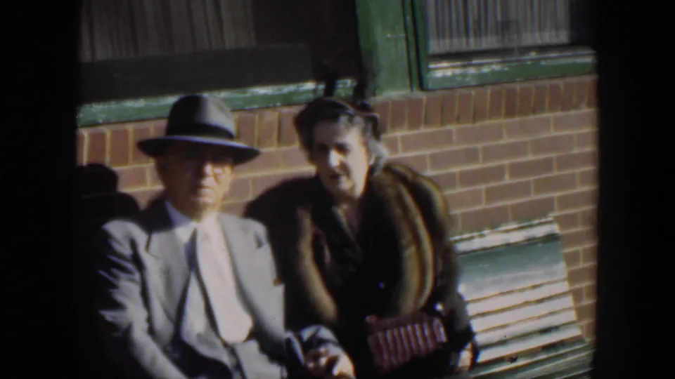 The seated couple looks toward the camera, framed by brickwork, green trim, and strong side light in the home-movie shot.