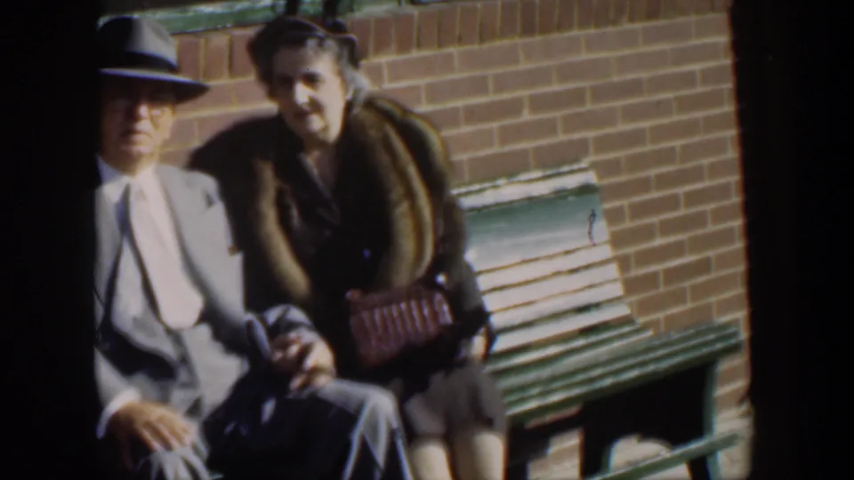 The couple remains seated against a brick wall, with the man in a pale suit and hat and the woman holding a patterned handbag.