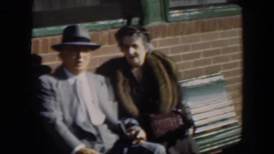 A mid-century home-movie frame shows the seated couple in warm light, with the woman leaning slightly toward the man on the green bench.