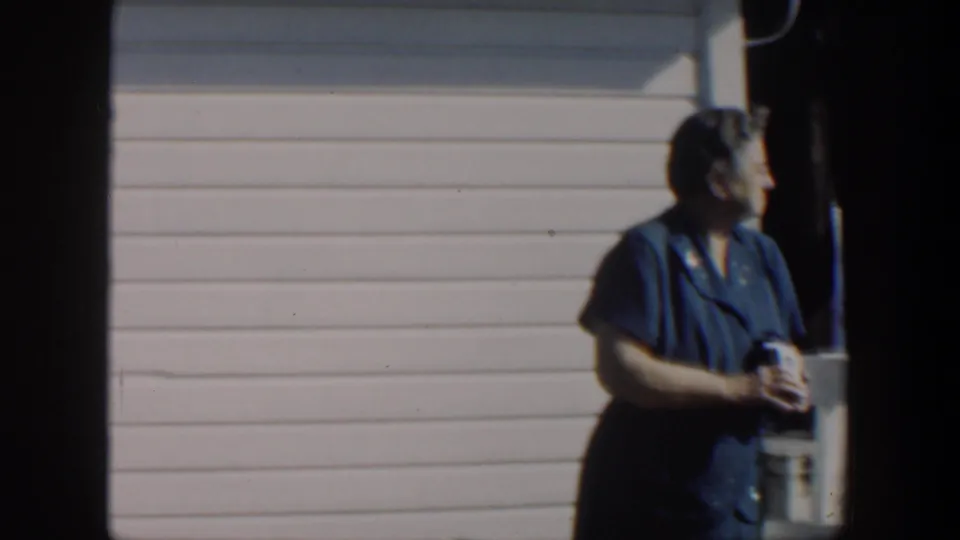 1951 Cuba 8mm frame of an adult woman in a blue dress holding a small object beside pier posts and pale siding.