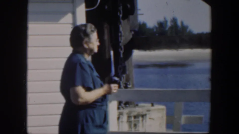 1951 Cuba 8mm frame of an adult woman beside a pier railing, with water, shoreline vegetation, and structural posts behind her.