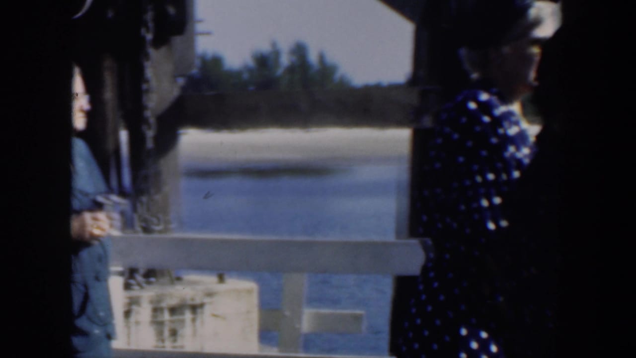 In 1951 Cuba footage, adult women in patterned dresses and hats stand by a waterside railing in bright sun.