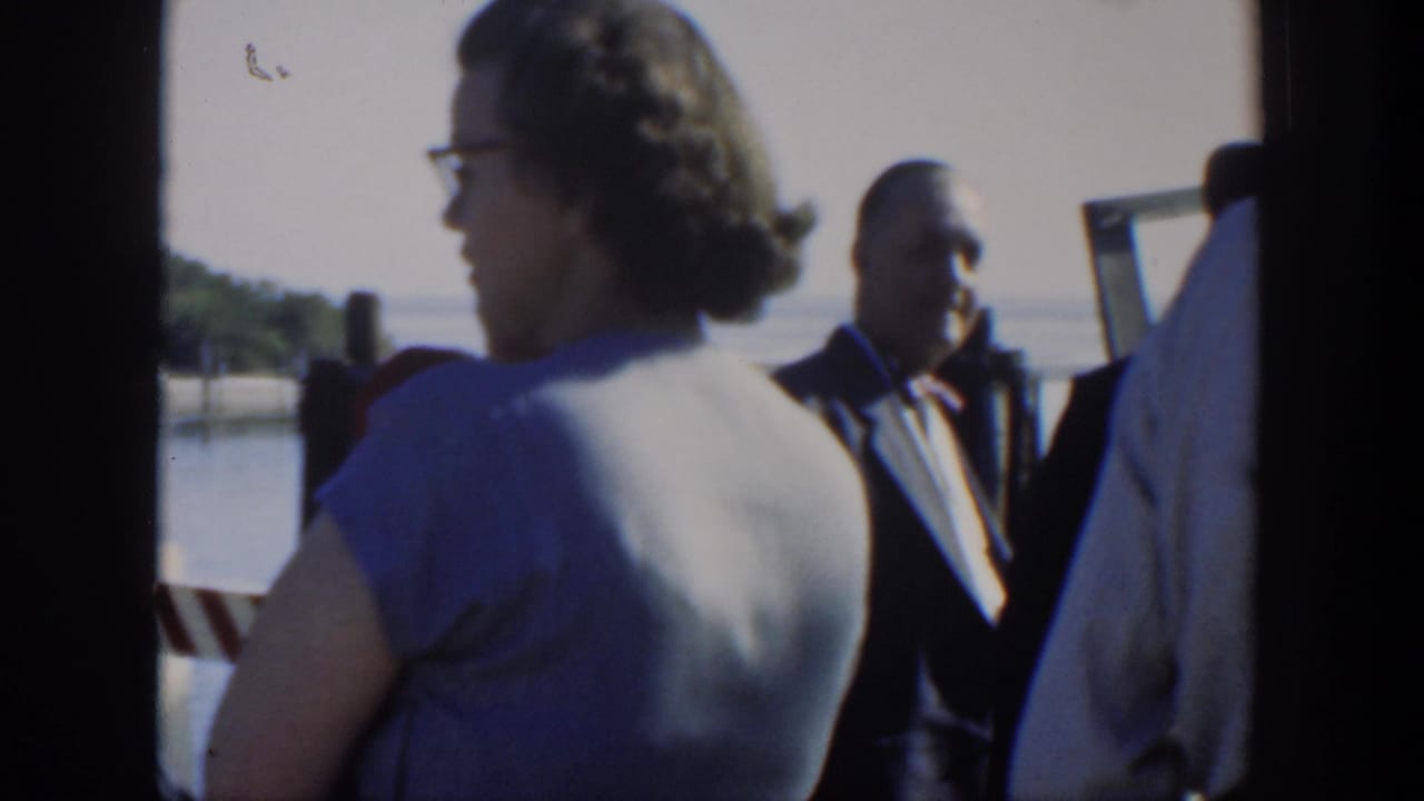 Adults stand near a waterfront, including a woman in glasses and men in suits and hats.