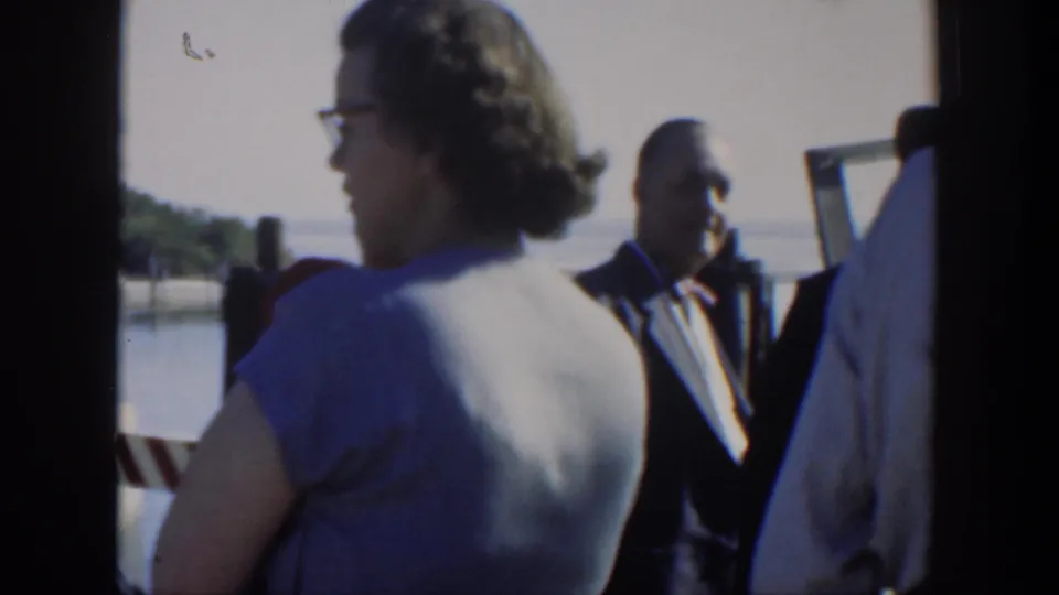 1951 Cuba home-movie frame showing adults gathered by a marina or waterfront, with a woman in glasses in the foreground and suited men behind her.
