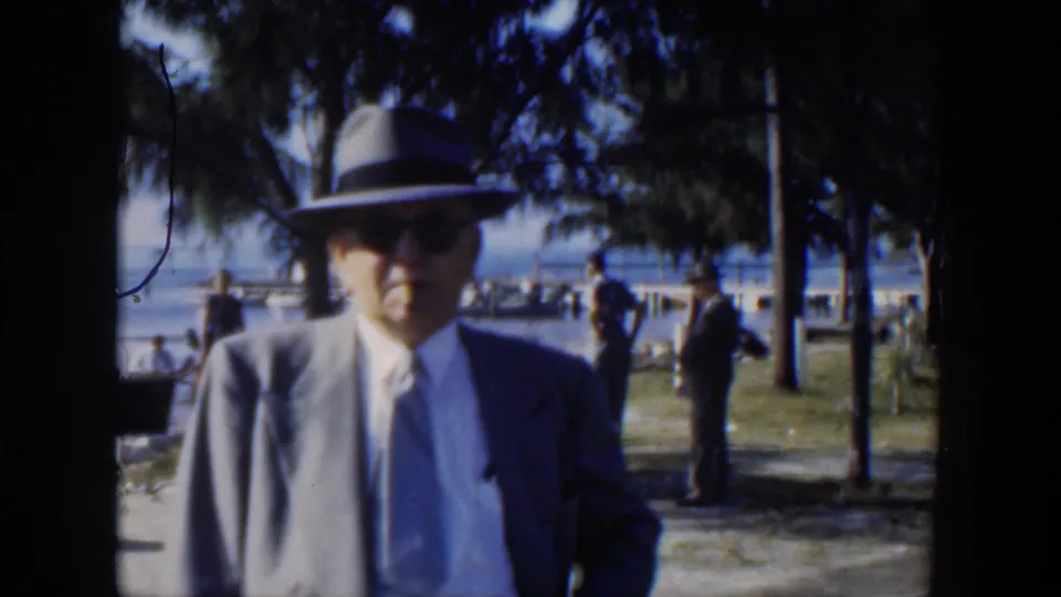 1951 Cuba home-movie frame of a suited man in a fedora facing the camera, with trees, grass, people, and water behind him.