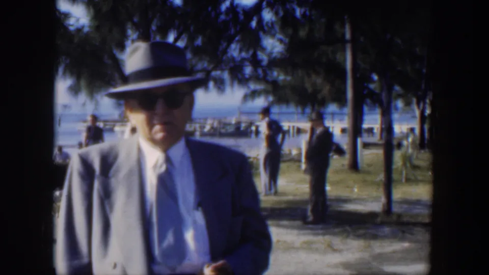 1951 Cuba frame showing a man in a fedora and suit near the camera, with trees, grass, waterfront railings, and scattered adults in the background.