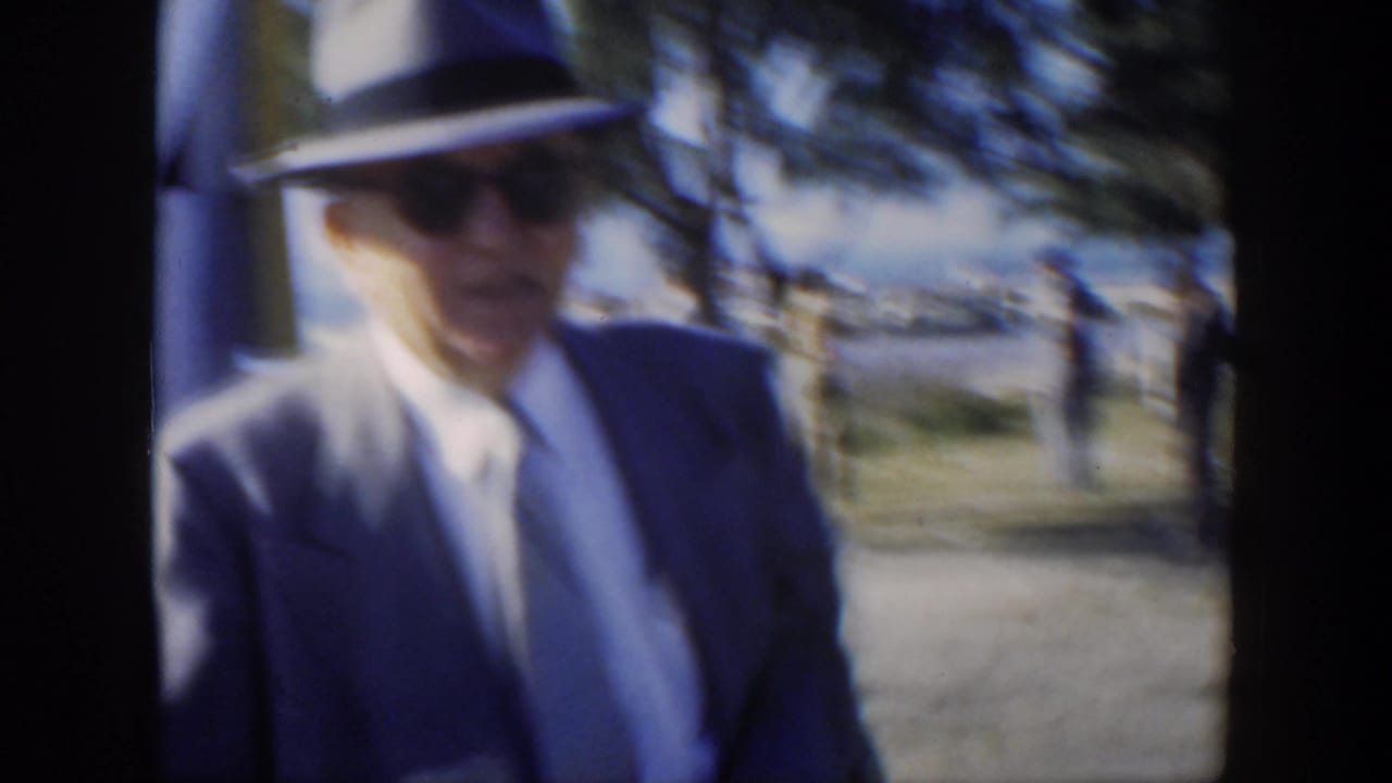 In 1951 Cuba, a well-dressed man in a light suit, fedora, tie, and sunglasses passes near the camera in a seaside park.