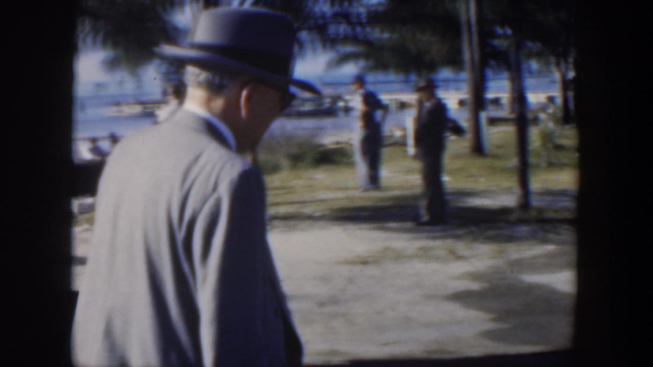 Man in fedora seen from behind walking along a park path with water beyond trees.