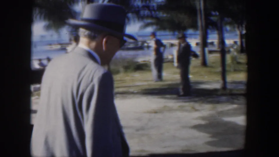 The man in a light suit and fedora walks away through a Cuban seaside park, with trees, path, pedestrians, and water visible beyond.