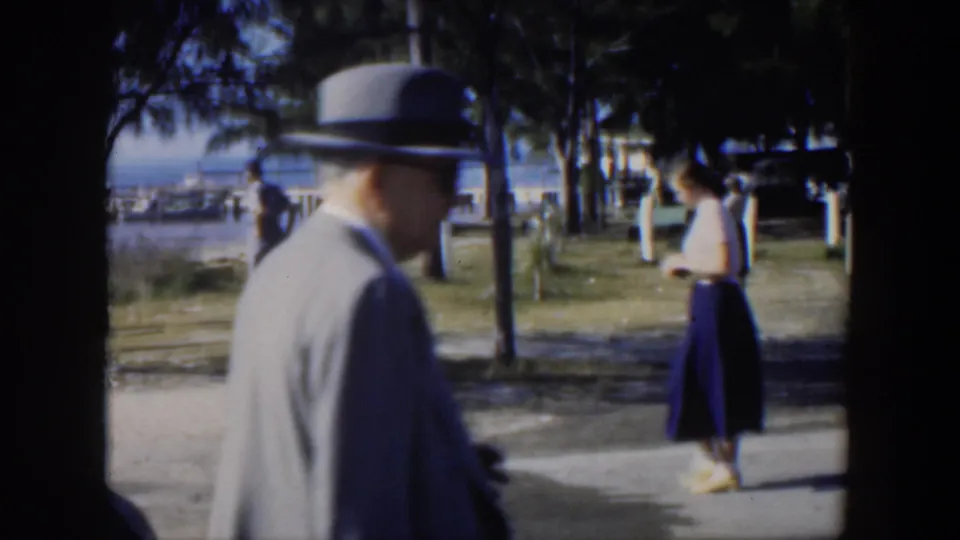A suited man in a fedora moves through a 1951 Cuba seaside park as a woman stands nearby and other figures appear in the distance.