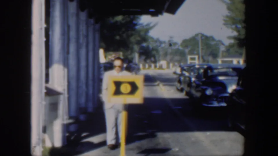A man in sunglasses and light clothing passes a yellow service sign while dark vintage cars are parked in a sunlit Cuba street scene.