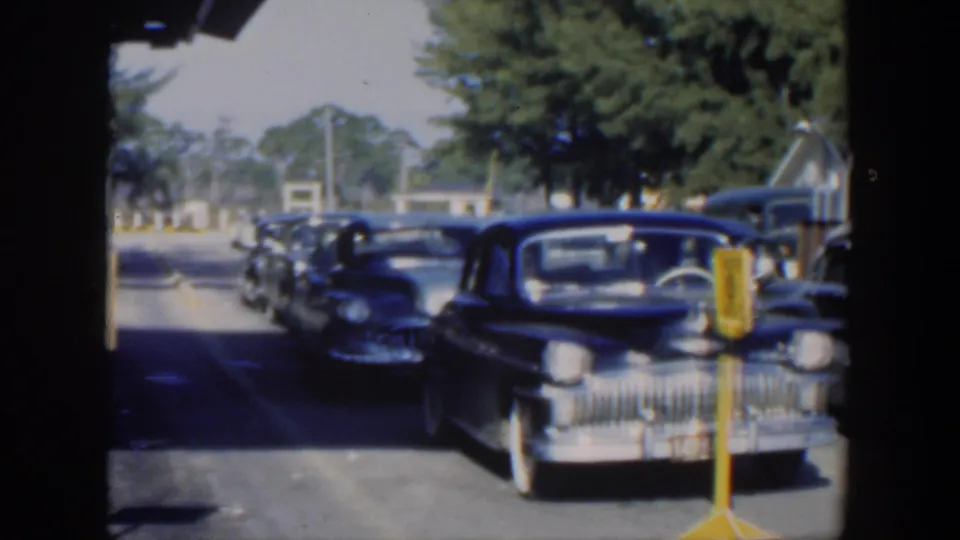 Rows of dark mid-century cars approach or sit near yellow service signs in a bright 1951 Cuba automotive setting.