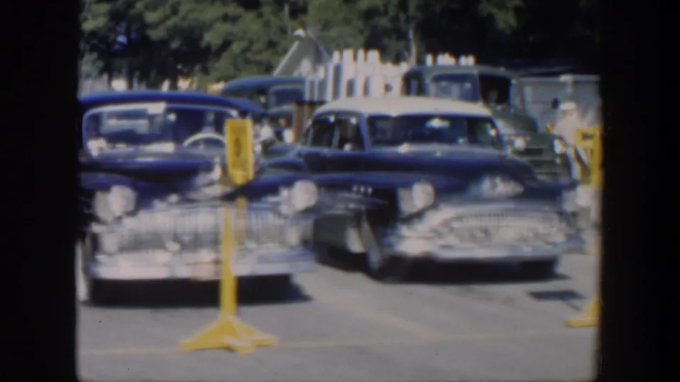 Front-facing dark American cars stand by yellow service signs, with more vehicles and roadside buildings visible in the 1951 Cuba frame.