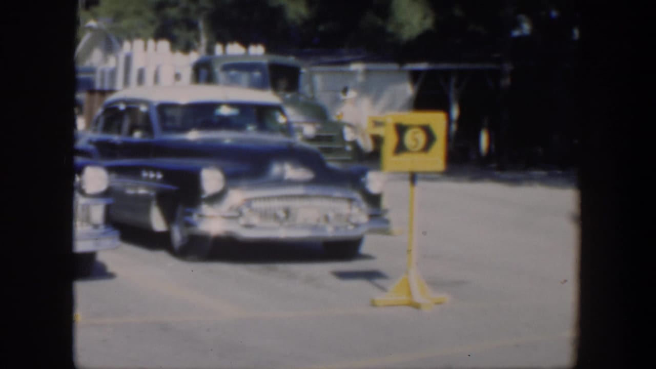 1951 Cuba home-movie frame showing a blue mid-century car approaching near a yellow numbered sign in a sunlit outdoor lot.