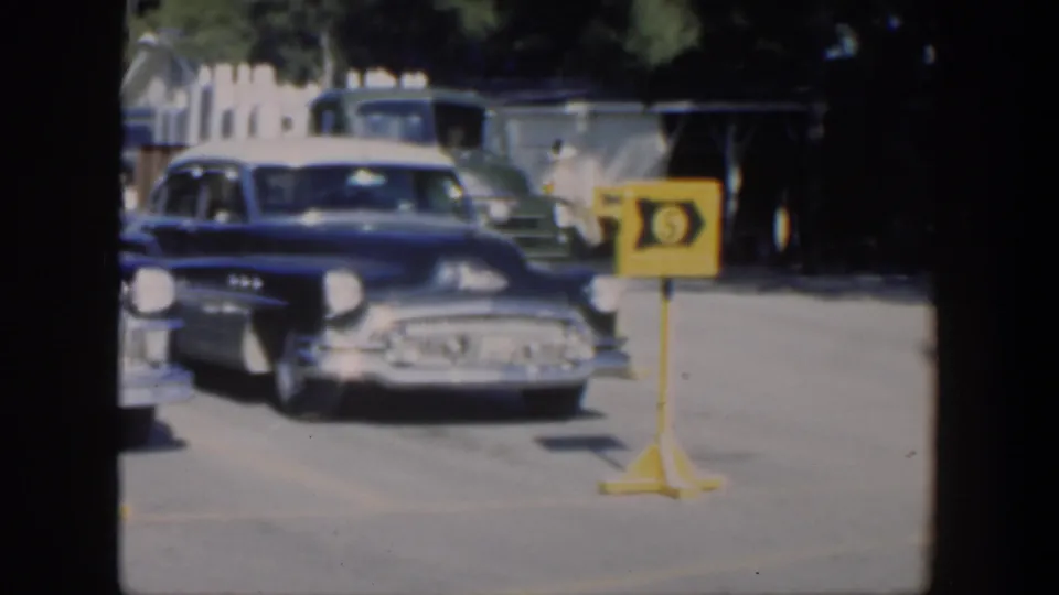 1951 Cuba home-movie frame showing a blue mid-century car approaching near a yellow numbered sign in a sunlit outdoor lot.